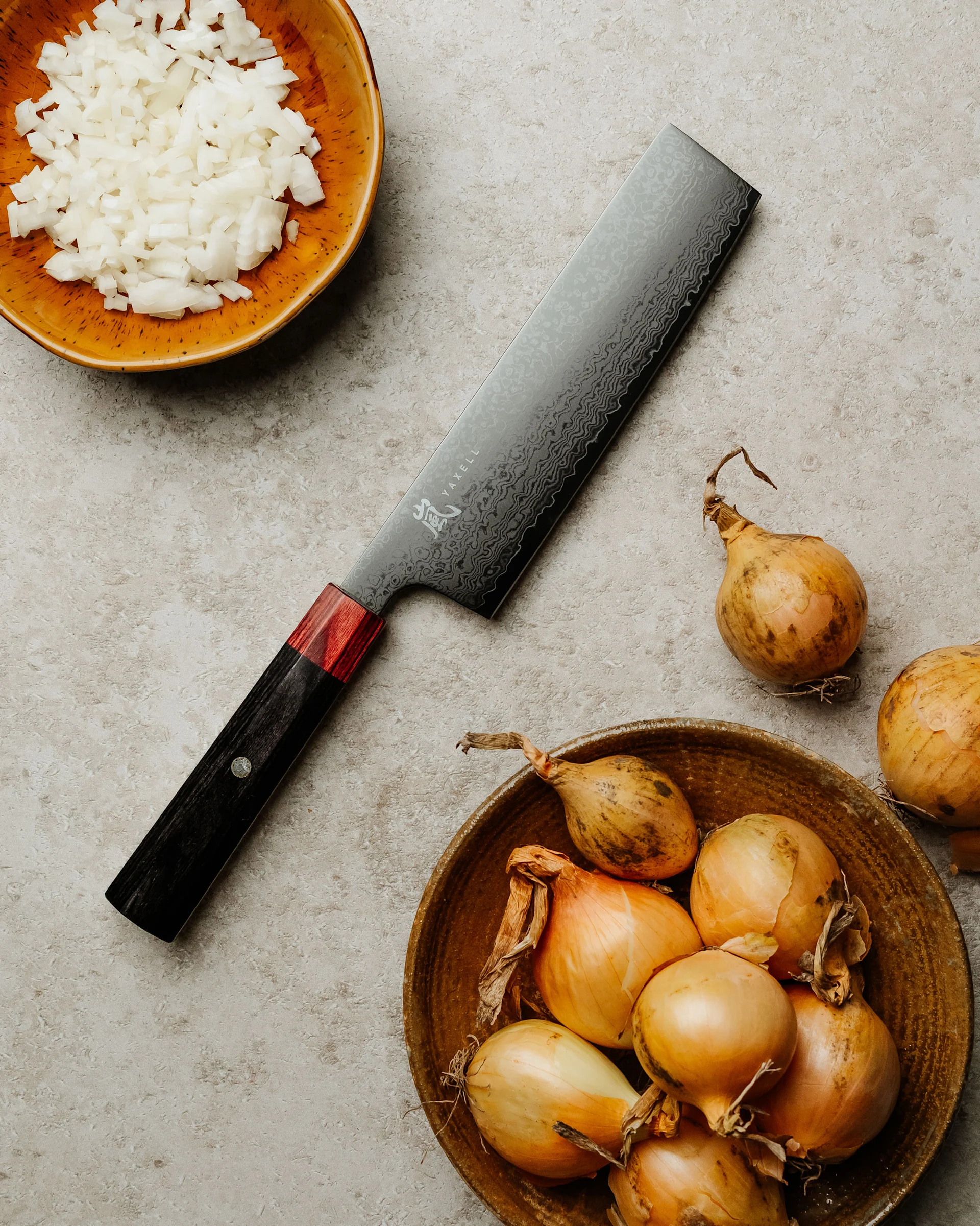 En kniv med mønstret blad og rødt/svart håndtak ligger ved siden av en skål med hakket løk og en skål med hele løk på en lys overflate.