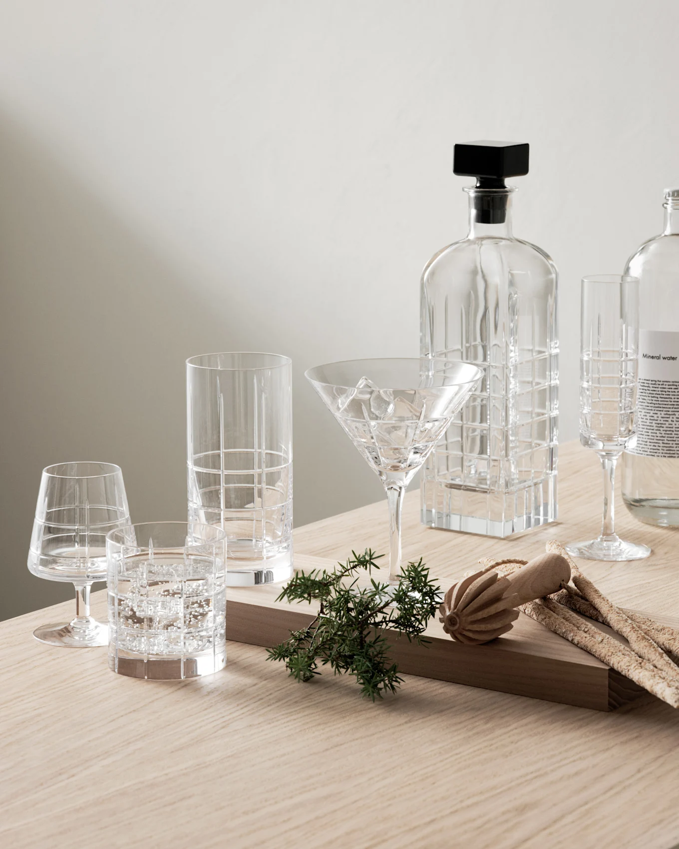 Klar glasskaraffel, ulike mønstrede glass og en mineralvannflaske med einer og grissini på et trebord.