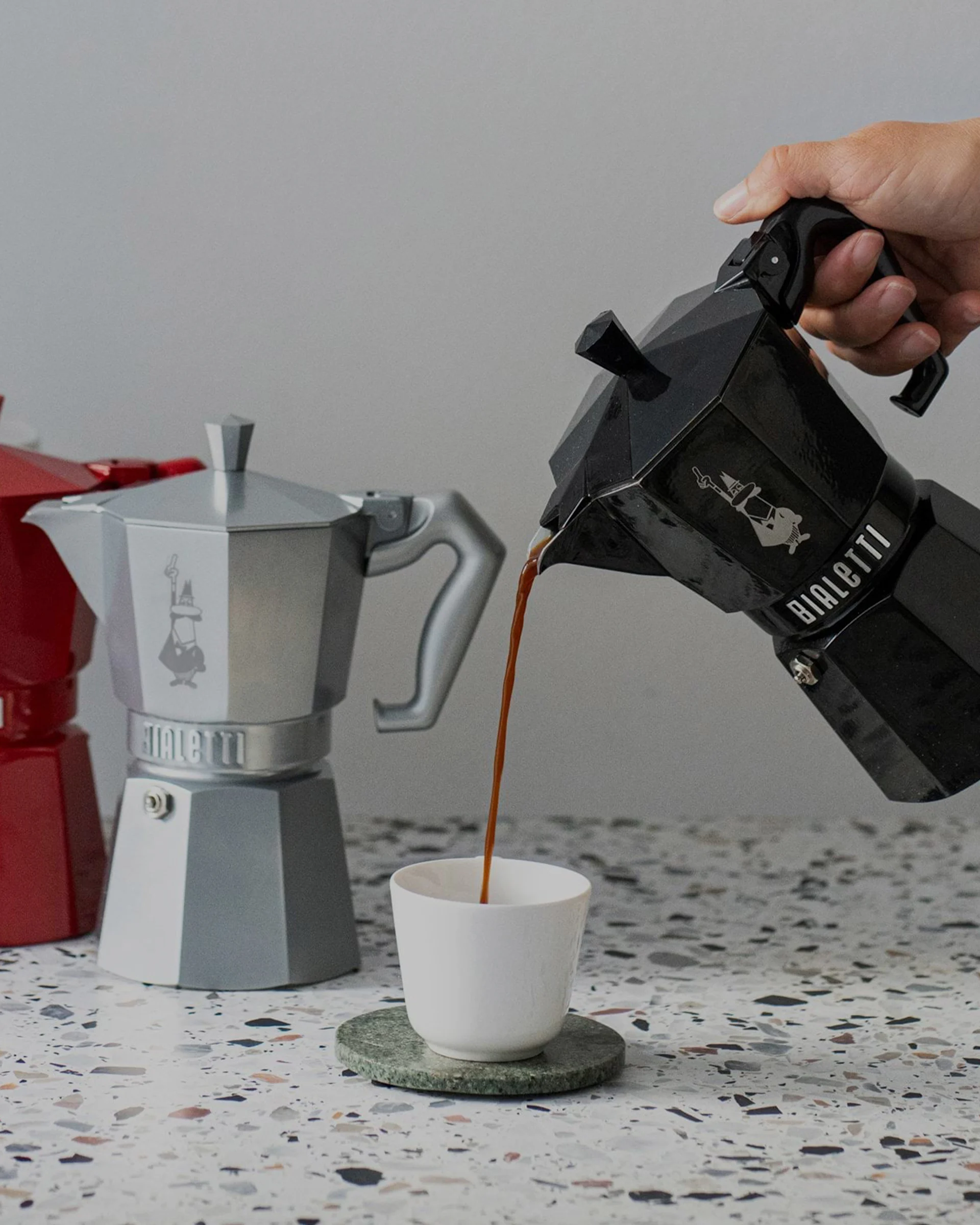 Hånd heller kaffe fra svart mokkakanne i hvit kopp, med sølv og rød mokkakanne i bakgrunnen på flekkete benk.