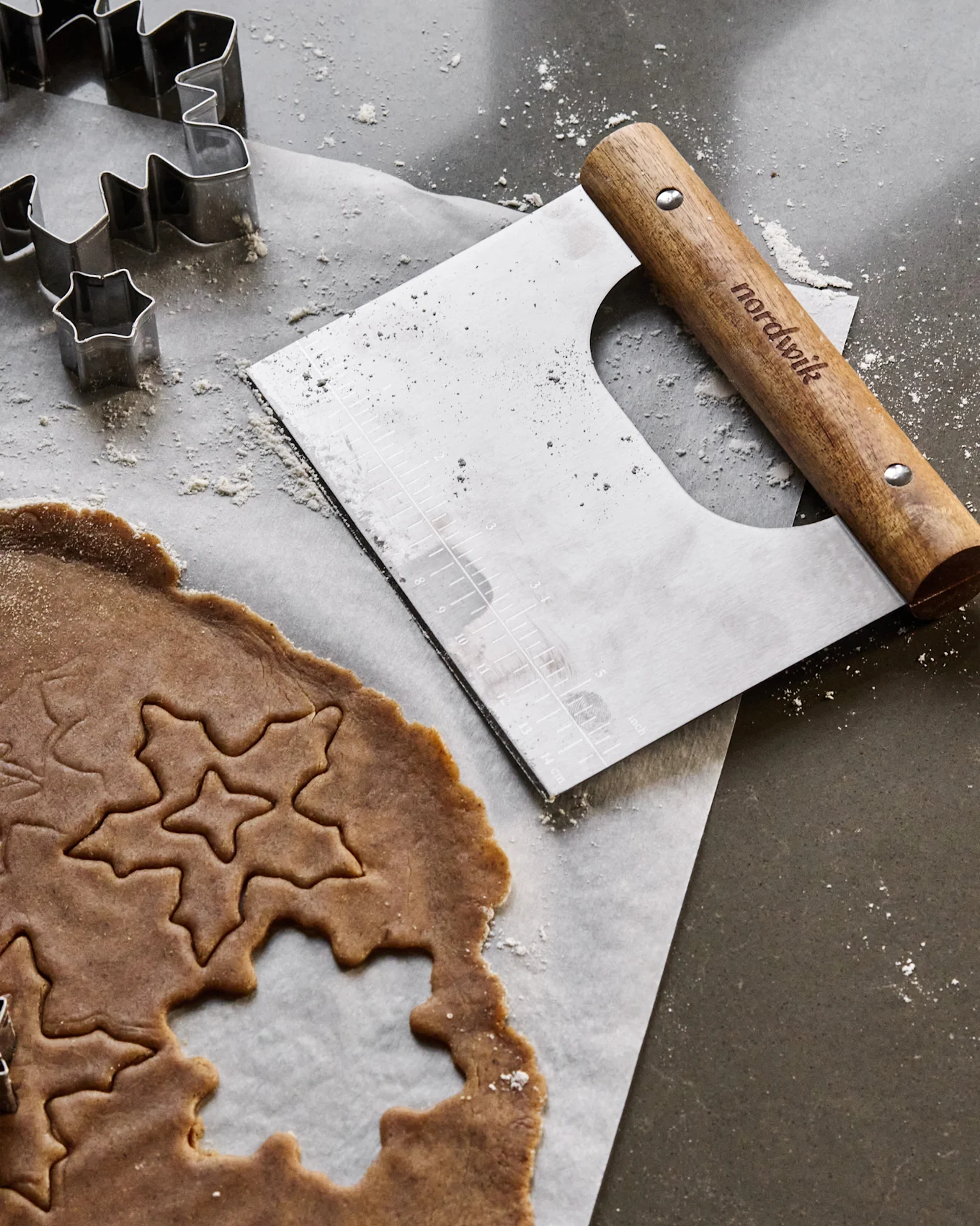 Pepperkakedeig med stjerne- og bladvutstansinger, metallformer og en deigskrape på en melet overflate.