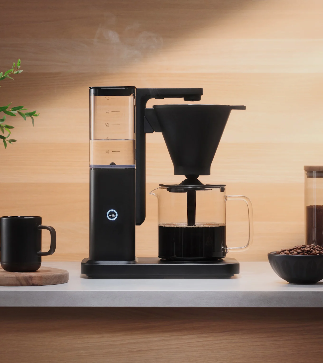 En svart kaffemaskin brygger kaffe i en glasskanne på en lys benk, med damp som stiger opp. En svart kopp og kaffebønner er i nærheten.