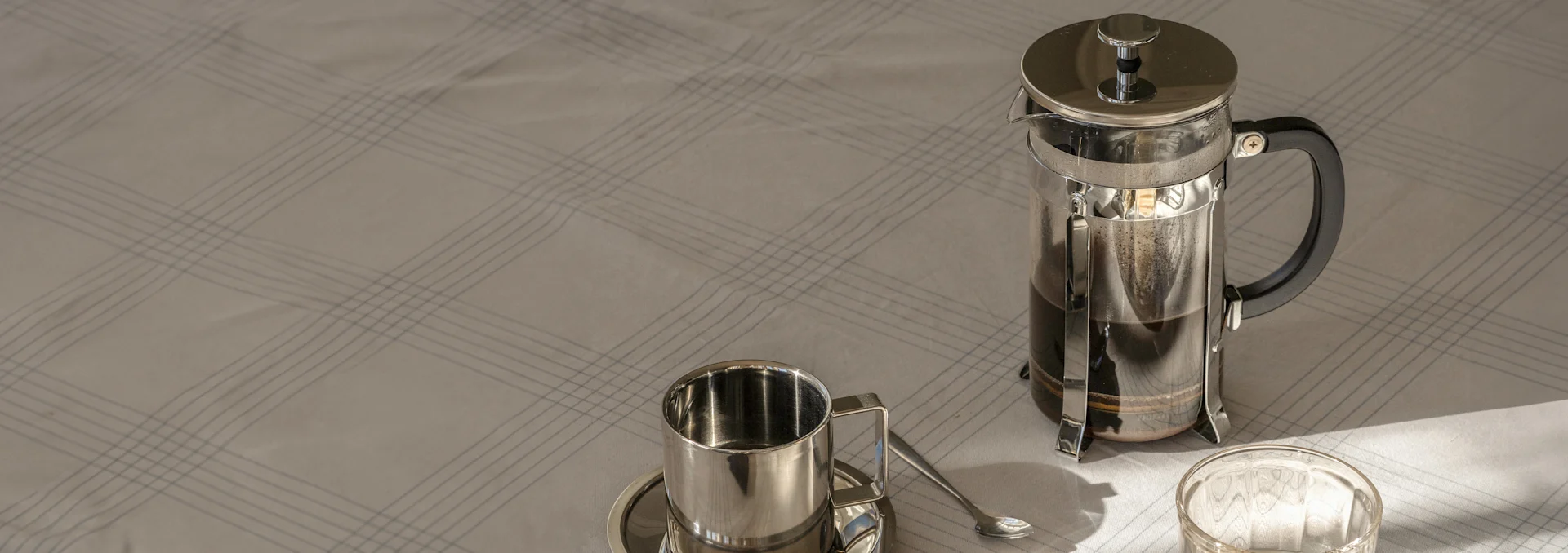French press med kaffe, en metallkopp og skål med skje på en lys, rutete duk i sollys.