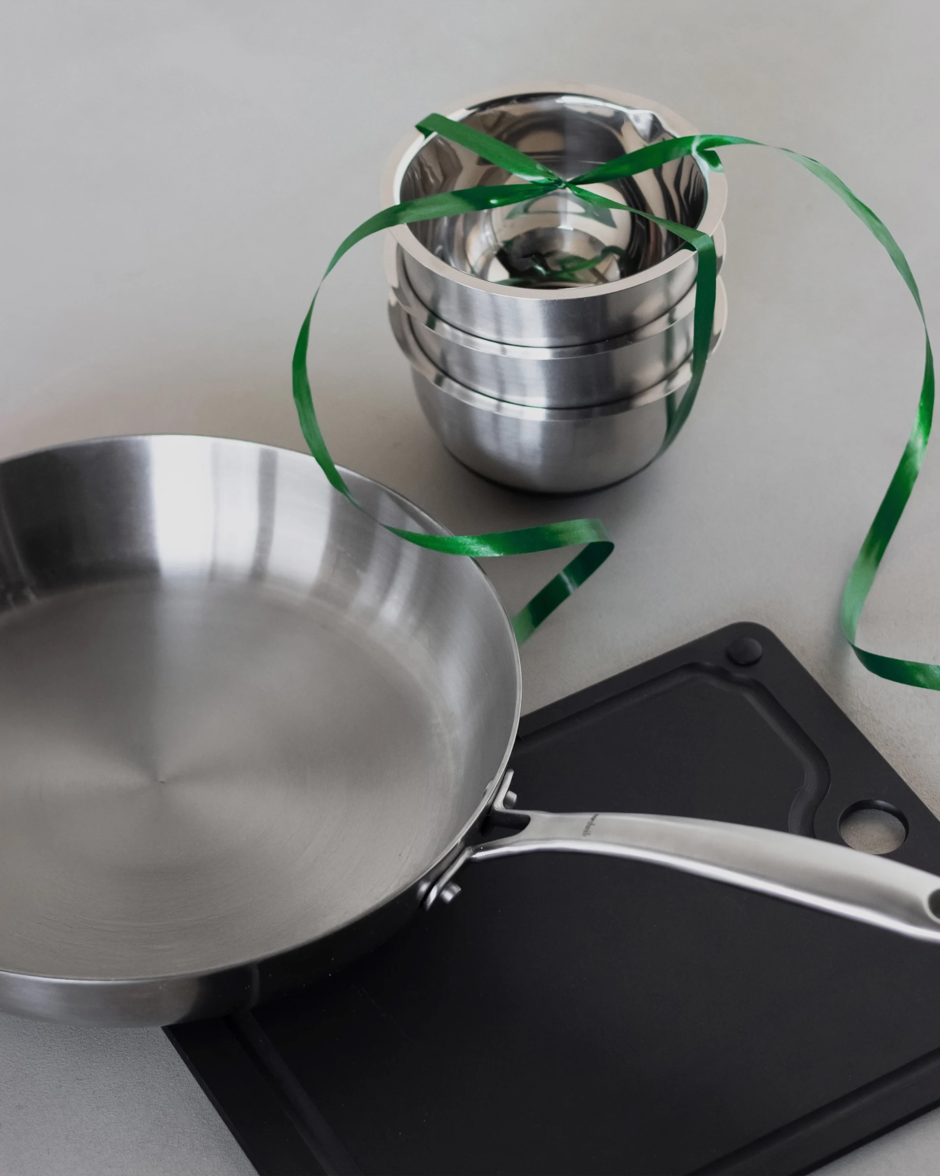 En stekepanne i rustfritt stål, en stabel metallboller bundet med et grønt bånd og et svart skjærebrett.