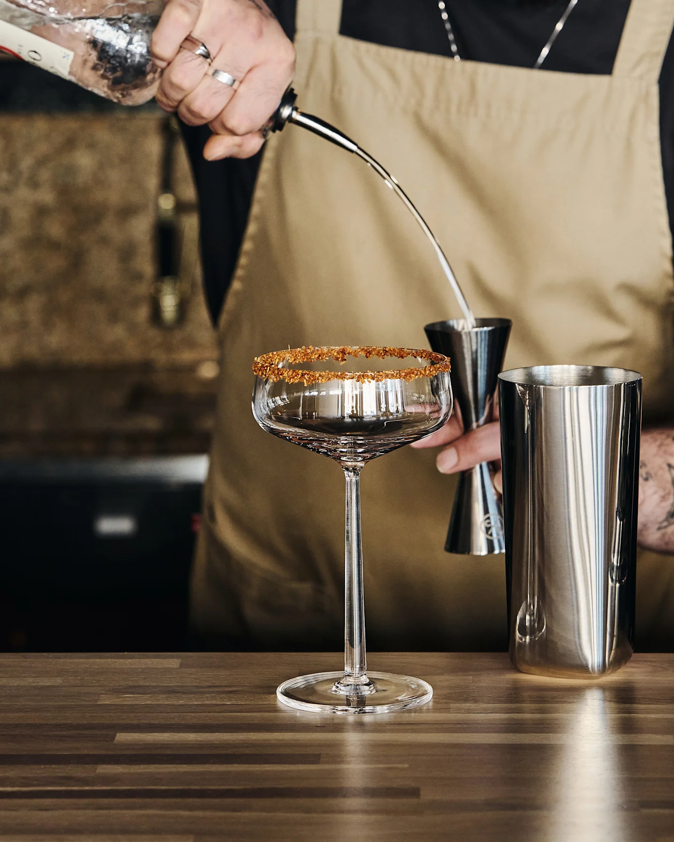 Bartender heller væske fra en flaske i et målebeger over en shaker, ved siden av et stettglass med sukkerkant.