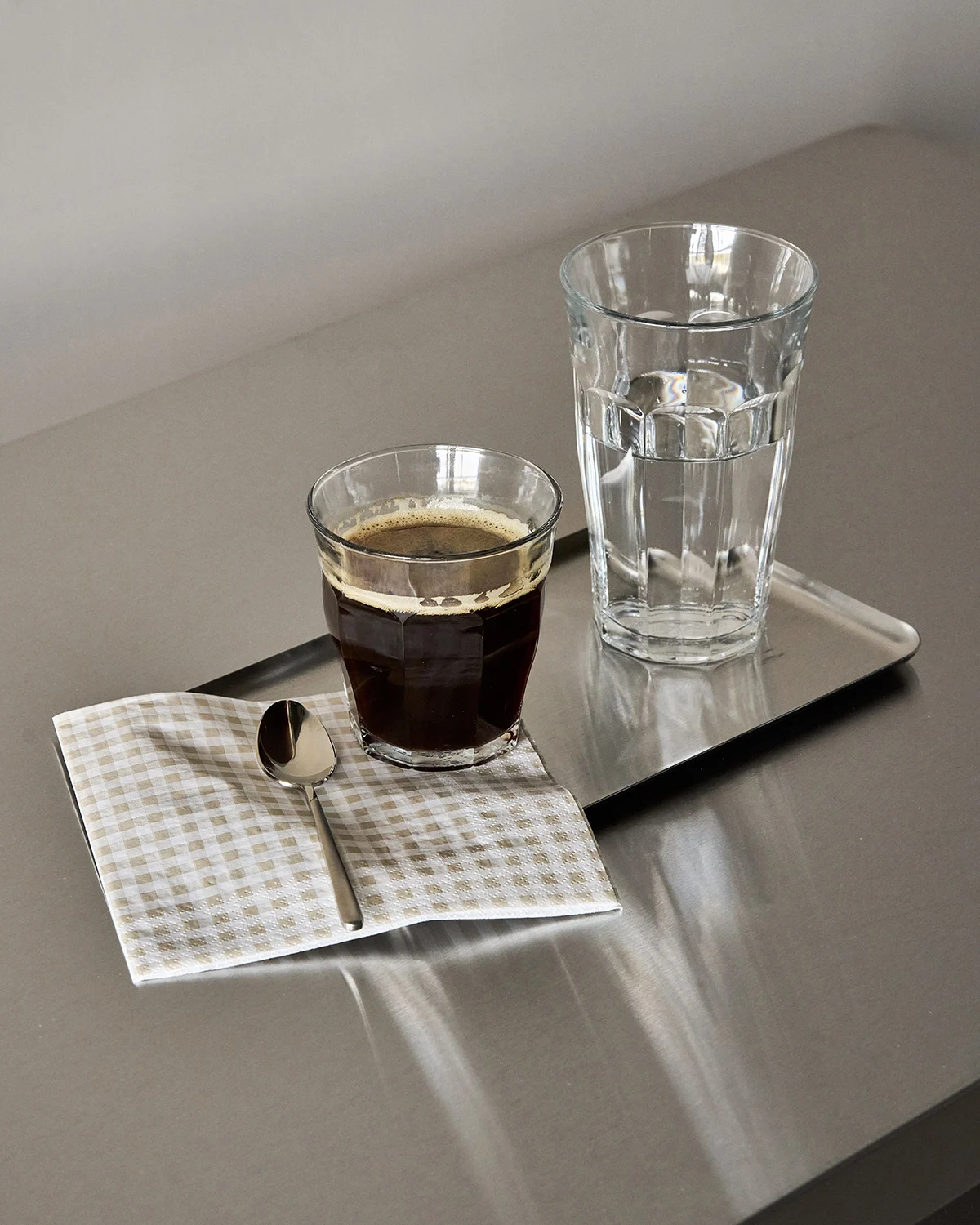 Glass sort kaffe, glass vann og en skje på en rutet serviett på et metallbrett.